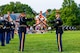 Two men in Army dark ceremonial uniforms are tossing brown rifles to each other while standing on a green lawn at an event.