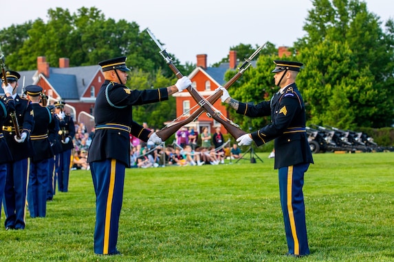 Two men in Army dark ceremonial uniforms are tossing brown rifles to each other while standing on a green lawn at an event.