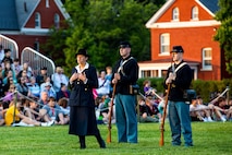 Three people (one woman and two men) are wearing Civil War-era clothing (the men are wearing Union Army uniforms, the woman is in a dark jack and skirt with dark hat), while delivering a dramatic presentation at an event on a green lawn.