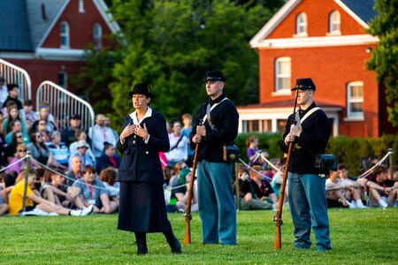 Three people (one woman and two men) are wearing Civil War-era clothing (the men are wearing Union Army uniforms, the woman is in a dark jack and skirt with dark hat), while delivering a dramatic presentation at an event on a green lawn.