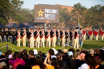 Soldiers dressed in Revolutionary War-era uniforms (dark blue coats with white and red trim, white pants and dark blue tri-cornered hats) are standing in rows in front of an audience at an event on a large green lawn. In the background is a large screen that reads Twilight Tattoo.