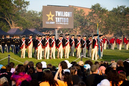 Soldiers dressed in Revolutionary War-era uniforms (dark blue coats with white and red trim, white pants and dark blue tri-cornered hats) are standing in rows in front of an audience at an event on a large green lawn. In the background is a large screen that reads Twilight Tattoo.