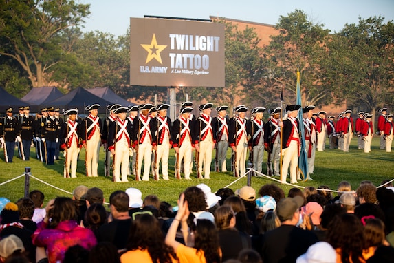 Soldiers dressed in Revolutionary War-era uniforms (dark blue coats with white and red trim, white pants and dark blue tri-cornered hats) are standing in rows in front of an audience at an event on a large green lawn. In the background is a large screen that reads Twilight Tattoo.