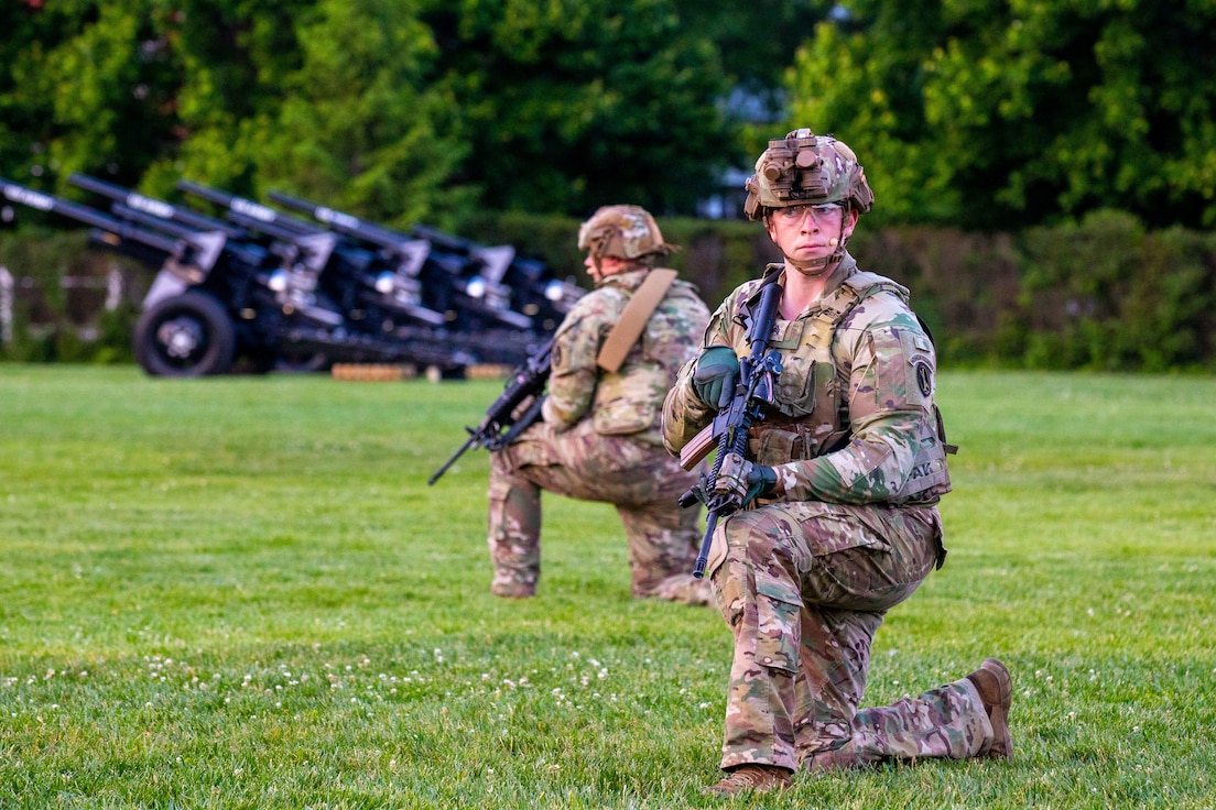 Army soldiers dressing in green camouflage uniforms and helmets are kneeling on a green lawn while looking around. They have black rifles in their hands, and there are black cannons in the background