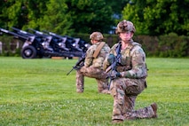 Army soldiers dressing in green camouflage uniforms and helmets are kneeling on a green lawn while looking around. They have black rifles in their hands, and there are black cannons in the background