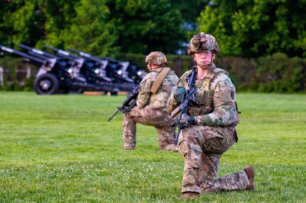 Army soldiers dressing in green camouflage uniforms and helmets are kneeling on a green lawn while looking around. They have black rifles in their hands, and there are black cannons in the background