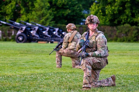 Army soldiers dressing in green camouflage uniforms and helmets are kneeling on a green lawn while looking around. They have black rifles in their hands, and there are black cannons in the background