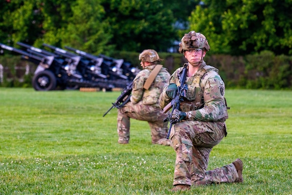 Army soldiers dressing in green camouflage uniforms and helmets are kneeling on a green lawn while looking around. They have black rifles in their hands, and there are black cannons in the background