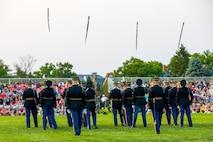 Soldiers dressed in dark Army service uniforms are facing away from the camera, and some are tossing rifles into the air. They are standing on a green lawn performing for an audience seated on metal bleachers in the distance.