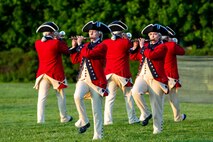 Soldiers dressed in Revolutionary War-era uniforms (red coats with blue and beige trim, white pants and dark blue tri-cornered hats) are marching while playing musical instruments on a green lawn.