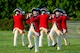 Soldiers dressed in Revolutionary War-era uniforms (red coats with blue and beige trim, white pants and dark blue tri-cornered hats) are marching while playing musical instruments on a green lawn.