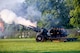 Black cannons resting on a green lawn are firing blanks at a ceremony. There is white and orange smoke coming out of their barrels.