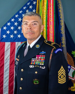 Latino man with black and grey hair wearing Army dark service uniform with many multicolored ribbons and gold-colored stripes on his sleeve is posing in front of the US flag and another flag that has dozens of ribbons coming from the top.