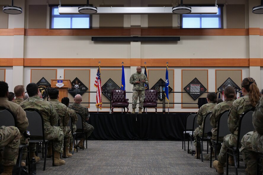 A man in military uniform stands center stage as he speaks to a crowd of military members in front of him.