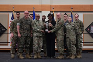 A group of uniformed military personnel stand on a stage huddled around a silver trophy for a group photo.