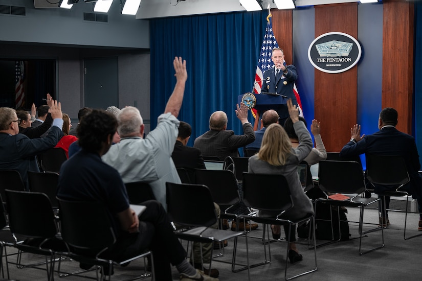 An Air Force general stands behind a lectern and points as members of a seated audience raise their hands.