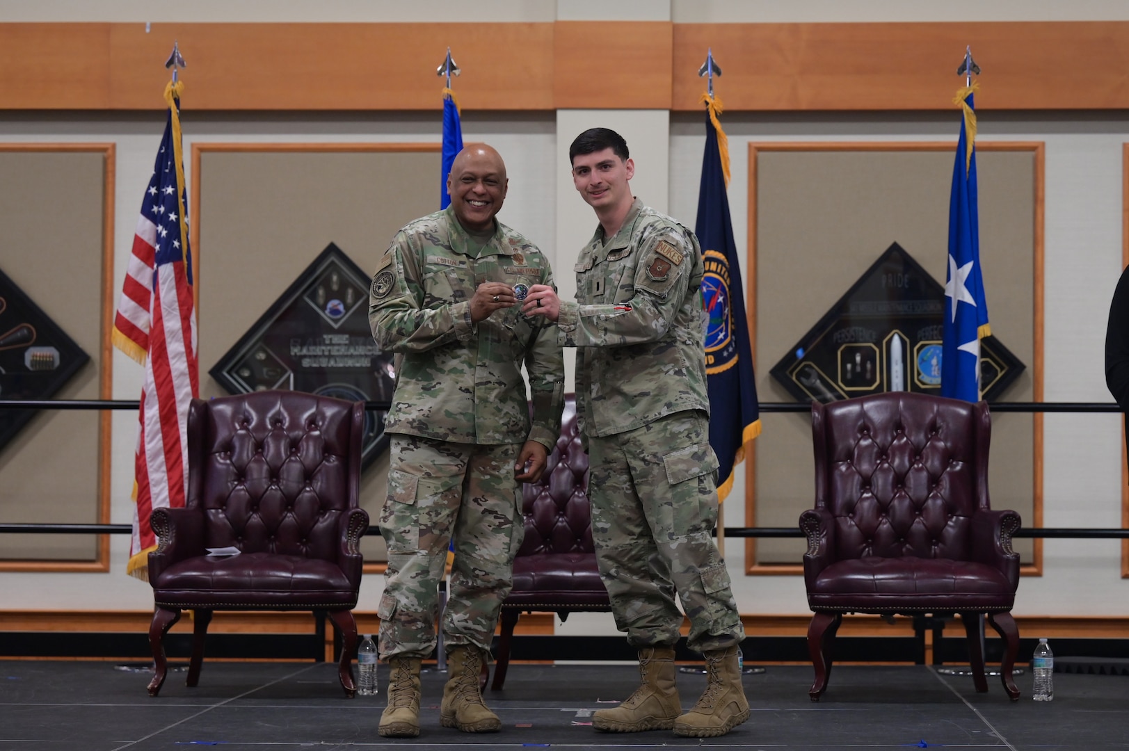 Two men in military uniform stand on stage as they hold a challenge coin together and smile toward the camera.