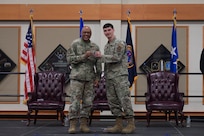 Two men in military uniform stand on stage as they hold a challenge coin together and smile toward the camera.