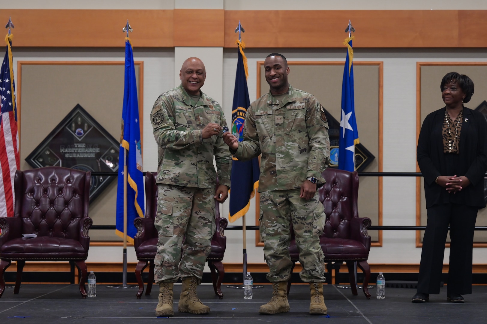 Two men in military uniform stand on stage as they hold a challenge coin together and smile toward the camera.