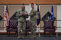 Two men in military uniform stand on stage as they hold a challenge coin together and smile toward the camera.