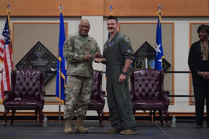 Two men in military uniform stand on stage as they hold a challenge coin together and smile toward the camera.