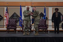 A man and woman in military uniform stand on stage as they hold a challenge coin together and smile toward the camera.