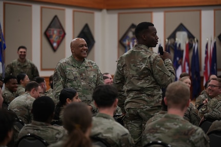 Two men in military uniform stand in a crowd of other seated military members.