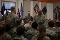 Two men in military uniform stand in a crowd of other seated military members.