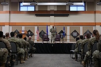 A man in military uniform stands center stage as he speaks to a crowd of military members in front of him.