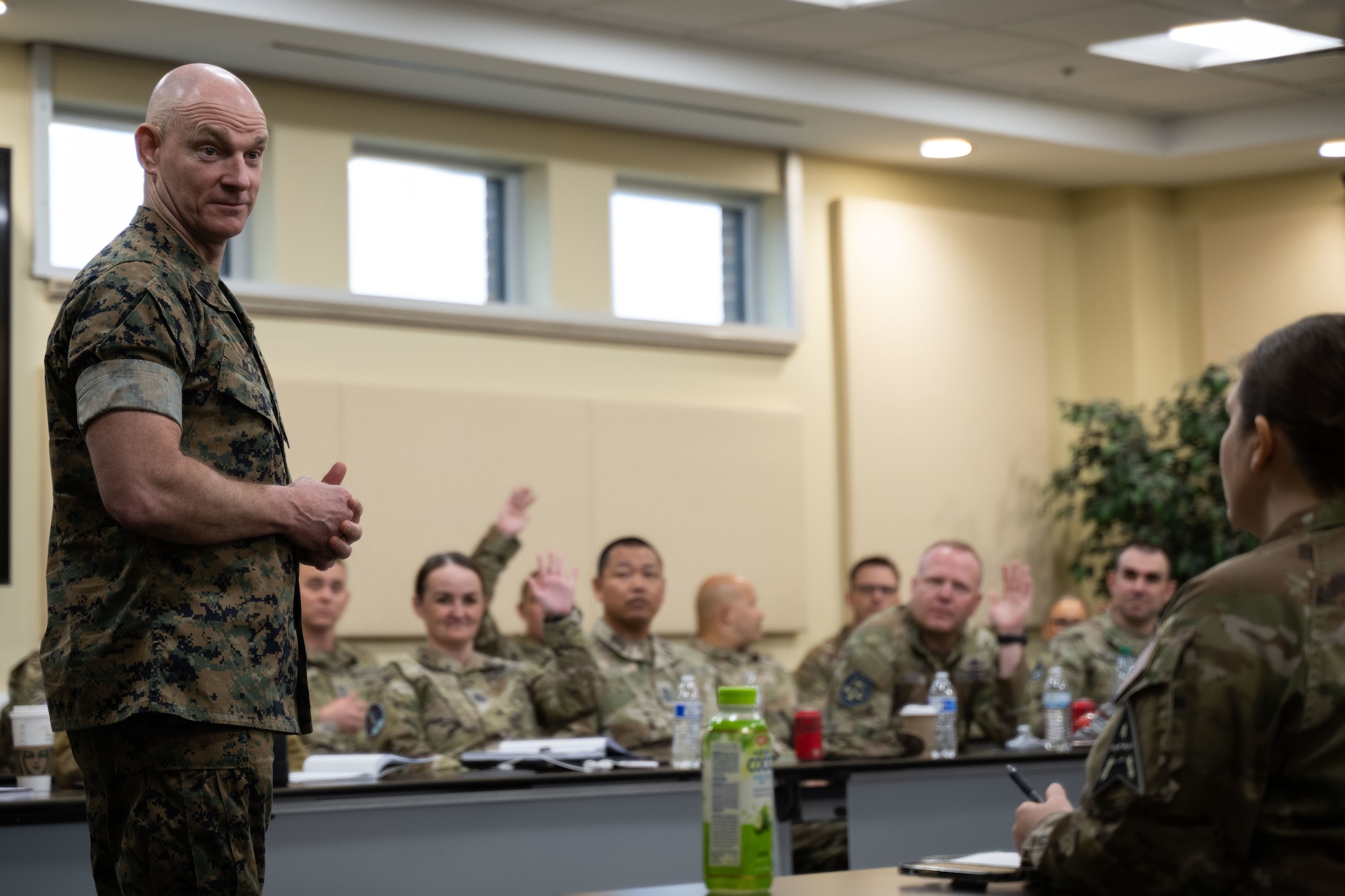 A military member stands and talks to other military members who are sitting in a classroom-like environment.