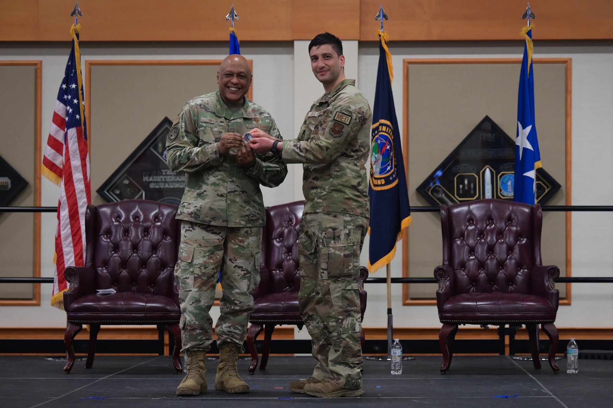 Two men in military uniform stand on stage as they hold a challenge coin together and smile toward the camera.