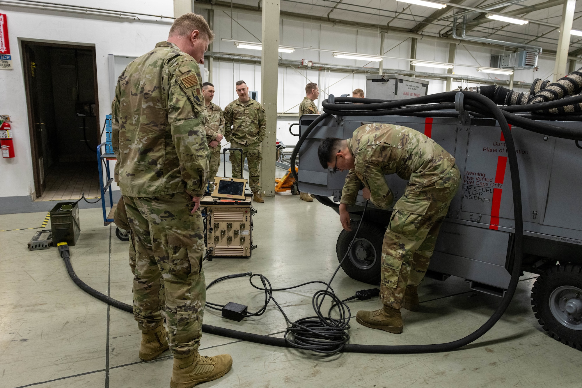 One man in military uniform shows another man in uniform how to plug a cable into a generator.