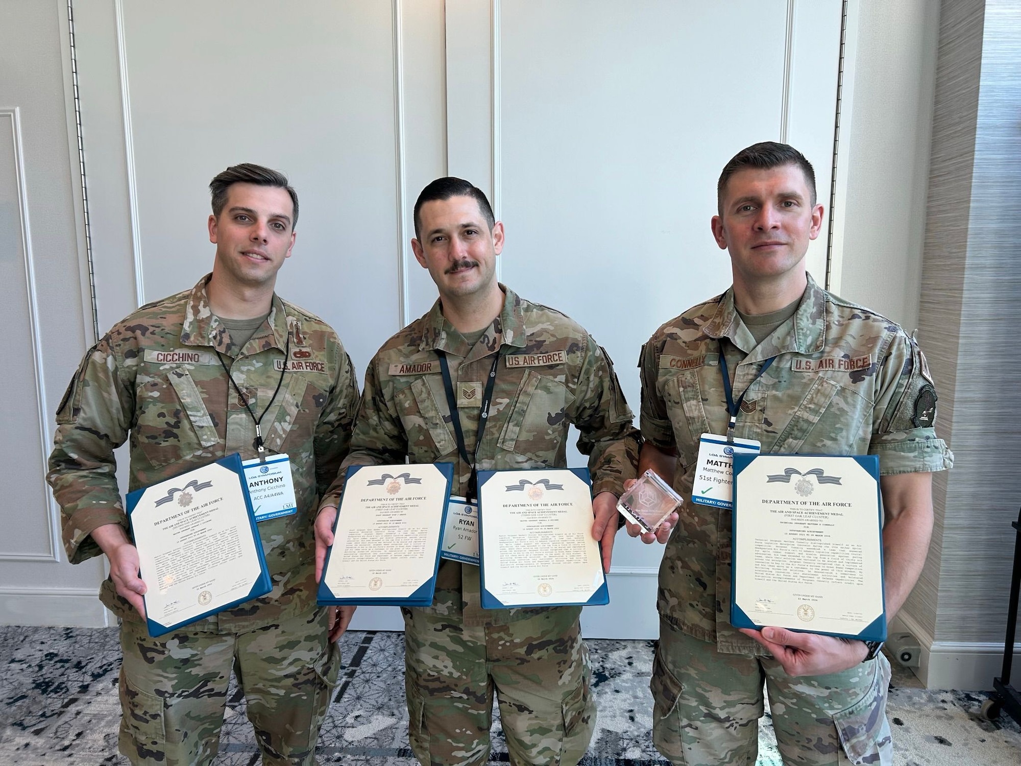Three men in military uniforms pose for a photo while holding certificates and a trophy.