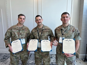 Three men in military uniforms pose for a photo while holding certificates and a trophy.