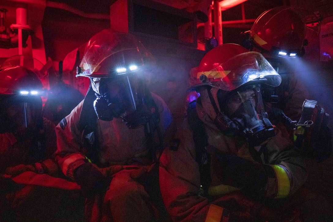 Sailors in firefighting gear use helmet lights while conducting training in a room with red lighting.