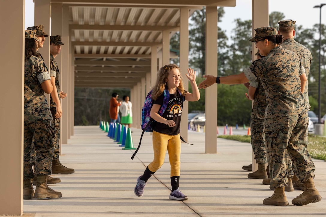 A Marine gives a student carrying a backpack a high-five outside a school. Other Marines stand in lines on both sides of the student.