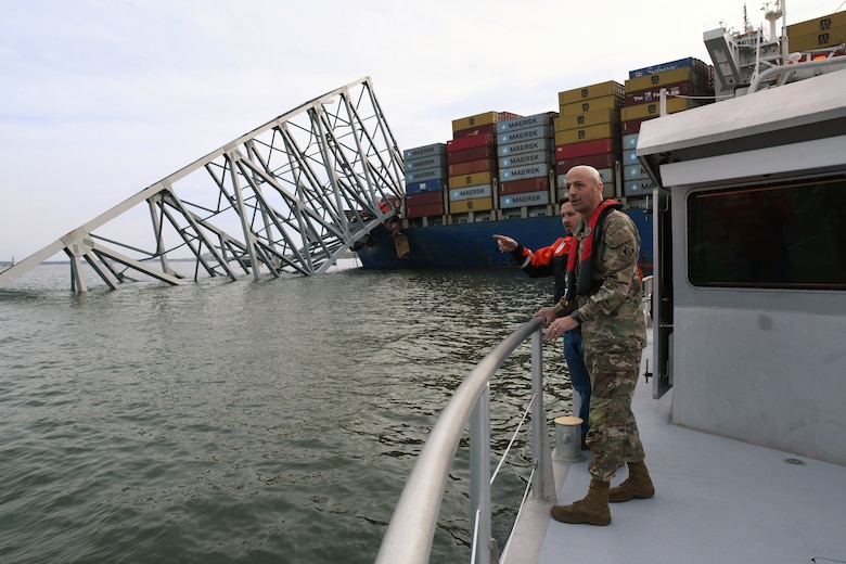 Two men on a white boat in a dark gray river overlook a bridge collapse with a blue cargo ship in the background.