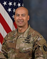 White man with bald head wearing Army green camouflage uniform posing in front of US flag