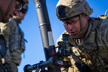 Two soldiers dressed in green Army combat uniforms with helmets and protective goggles are looking at a mortar making adjustments. The mortar is like a grey pipe that is pointing mostly upward but at a slight angle.