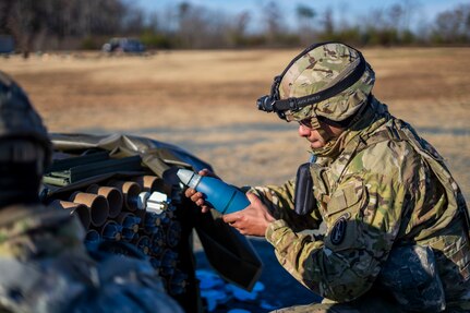 A soldier dressed in green Army combat camouflage uniform is holding a mortar round in his hands. The round is shaped like a rocket with a pointed tip, and it is blue-green in color. He's squatting in a field that has light brown grass.