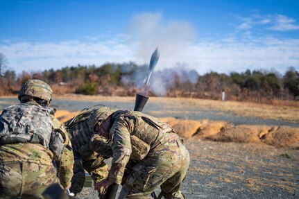 Soldiers dressed in green Army combat uniforms with helmets are huddled around a mortar as it is firing a round into the air. There is white smoke and the round is seen blasting out.