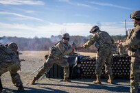 Soldiers dressed in green Army combat uniforms with helmets are standing around a mortar. One soldier is holding a round, which looks like a small missle, and he is about to drop it into the mortar to fire it.