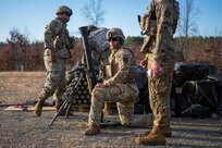 Soldiers dressed in green Army combat uniforms with helmets are huddled around a mortar, and one soldier is kneeling. The ground is grey gravel and sand.