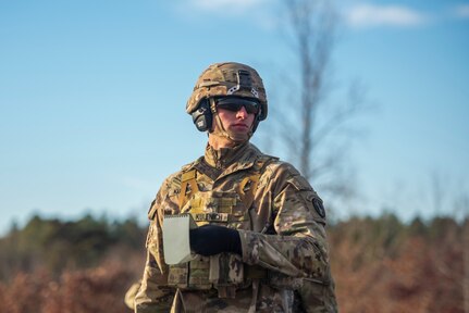 A soldier dressed in Army green combat camouflage and helmet with sunglasses and black gloves on is looking off to the side while holding a notepad in his had. There are trees and shrubs in the background.