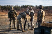 Soldiers dressed in green Army combat uniforms with helmets are standing around a mortar, about to load it with a round of ammunition.