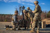 Soldiers dressed in green Army combat uniforms with helmets are huddled around a mortar and one Soldier is talking to one who is kneeling next to the mortar