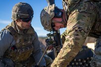 Two soldiers dressed in green Army combat uniforms with helmets are huddled around a mortar. They both have on sunglasses.