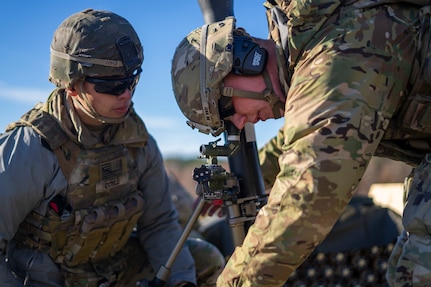 Two soldiers dressed in green Army combat uniforms with helmets are huddled around a mortar. They both have on sunglasses.