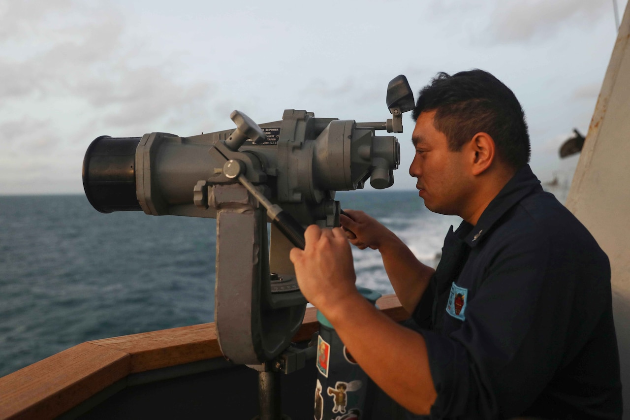 A service member in uniform looks through binoculars aboard a Navy vessel.