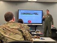 Man in flight suit does a hand gesture that appears to be a thumbs up pointed towards himself while presenting in front of a screen containing training materials with a military student in the foreground